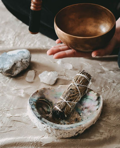  A hand holding a singing bowl next to crystals and to an abalone shell smudging bowl with a smudge stick inside. These materials are used for chakra healing, stress relief, or mindfulness rituals.