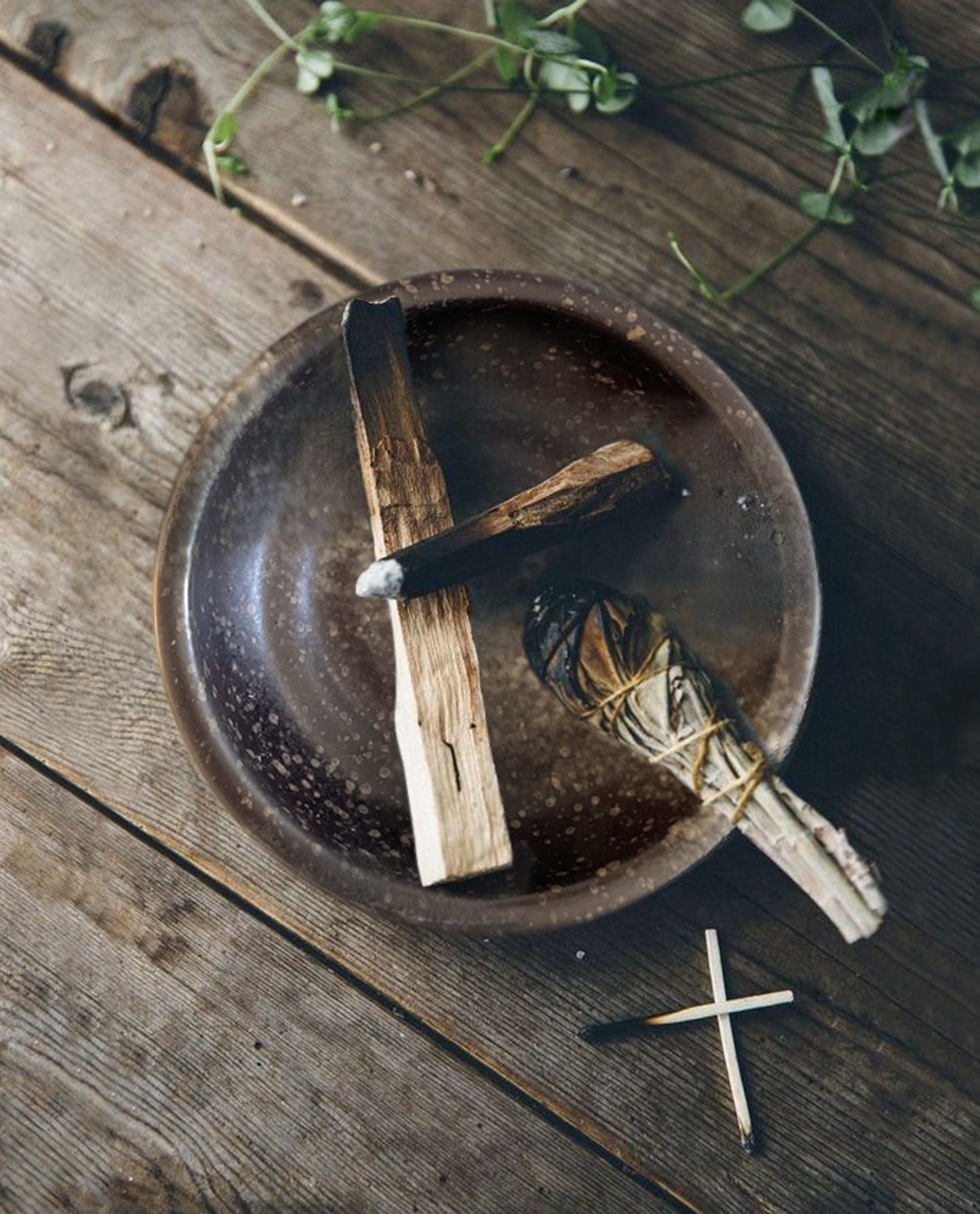  A smudging bowl with two palo santos and a smudge stick inside. The brown speckled ceramic smudging bowl is ready to be used to catch the ash produced during the smudging ritual.